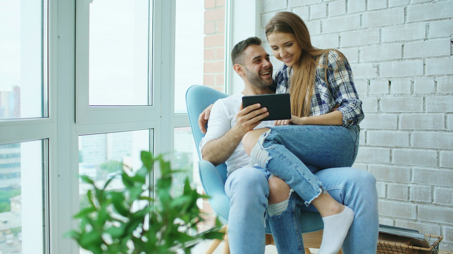 Couple laughing together while looking at a tablet in a modern living room.